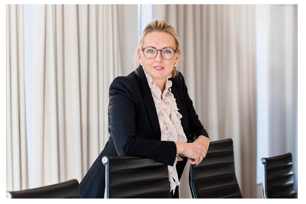 Kvinna i affärskläder lutar sig mot en stol i ett kontorsutrymme med gardiner i bakgrunden