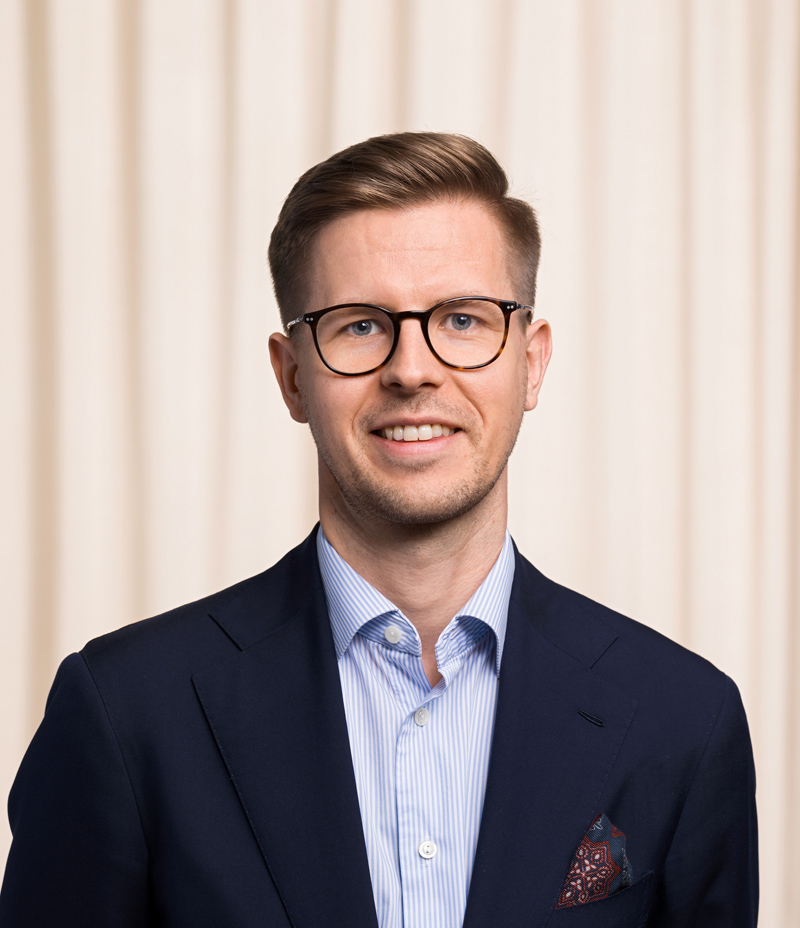 Man med glasögon och kort hår, klädd i mörk kostym och ljusblå skjorta, står framför en beige bakgrund