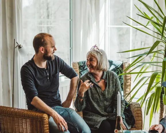 En man och en äldre kvinna sitter och samtalar i ett solbelyst rum med gröna växter i bakgrunden