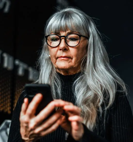 Äldre kvinna med grått hår och glasögon som använder en smartphone, klädd i svart tröja, inomhusmiljö