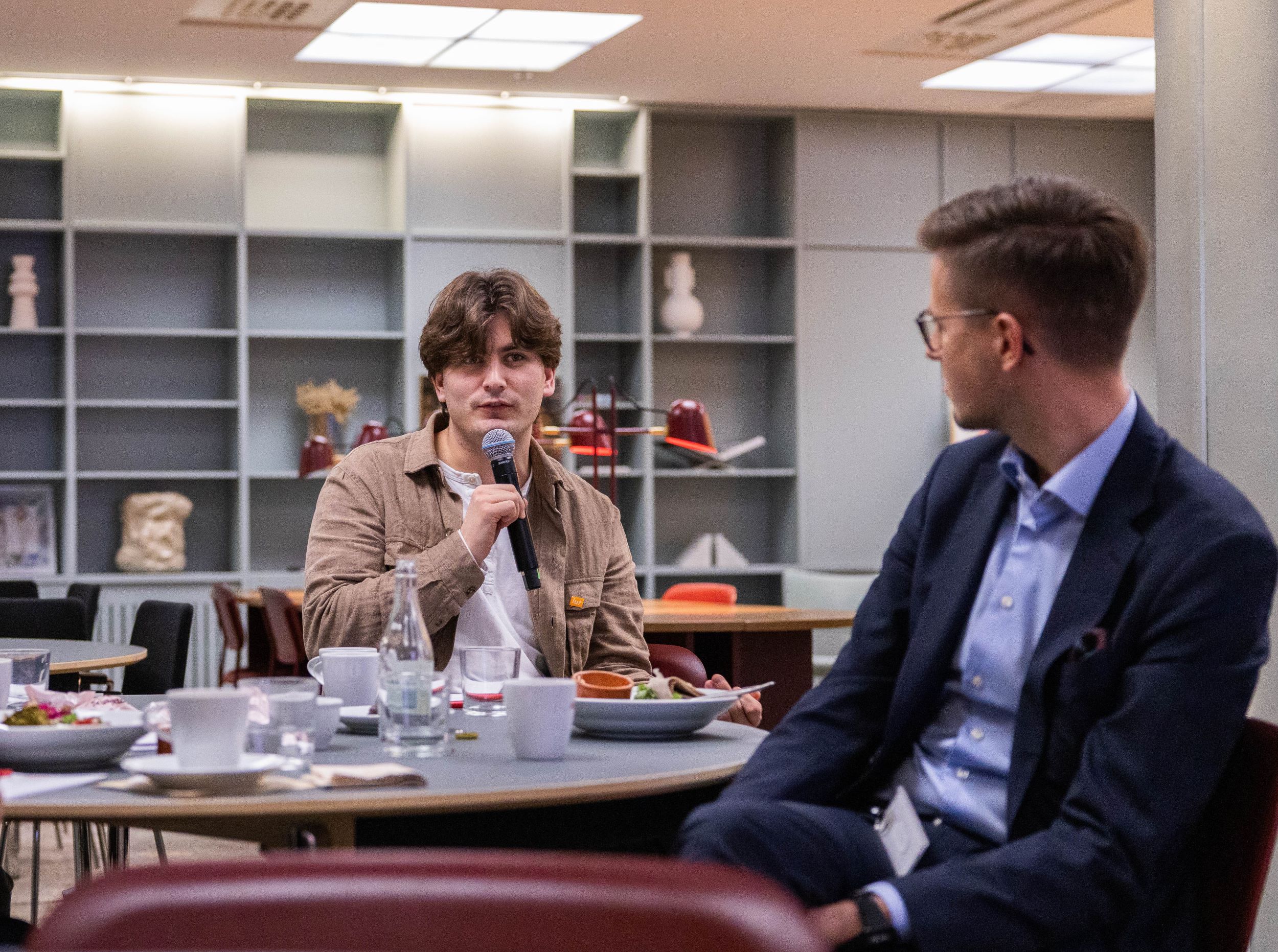 Personer i samtal vid en konferens eller affärsmöte, en man talar i mikrofon medan en annan man lyssnar, omgivna av bord med mat och dryck på ett modernt kontor