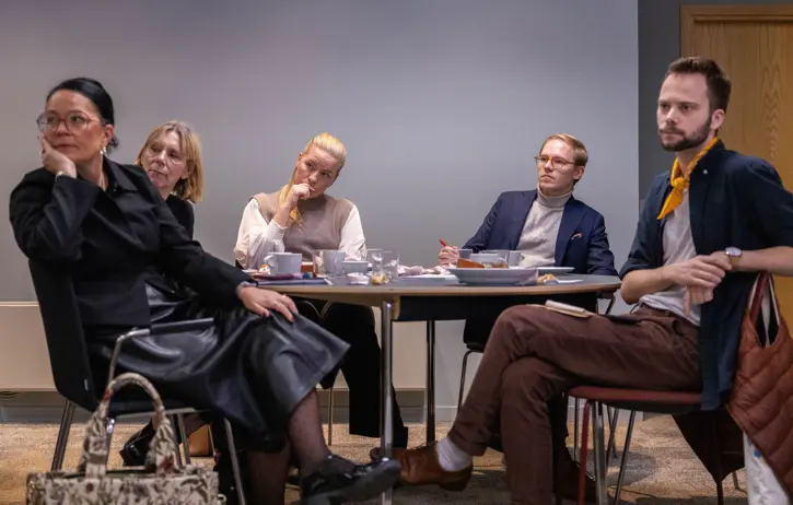Grupp människor deltar i ett möte runt ett bord, några med papper och kaffe framför sig, i ett kontorsliknande rum