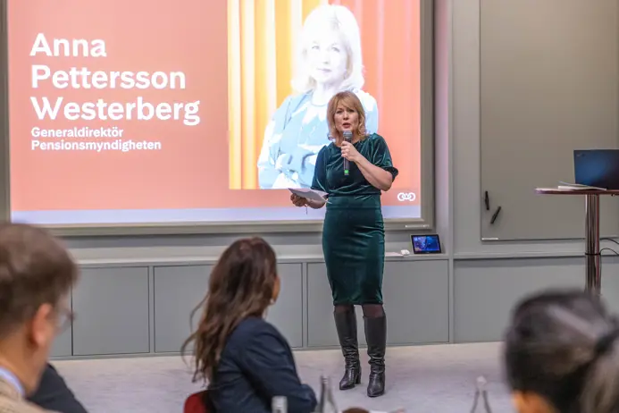 Kvinna i grön klänning talar på en konferens, framför en stor presentationsskärm. Texten på skärmen visar ett namn och en titel
