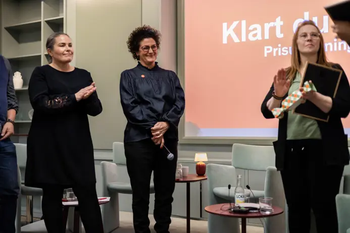 Tre personer står på en scen under en prisceremoni. En person håller en mikrofon, och en annan håller en present med ett randigt omslag. En presentation visas i bakgrunden