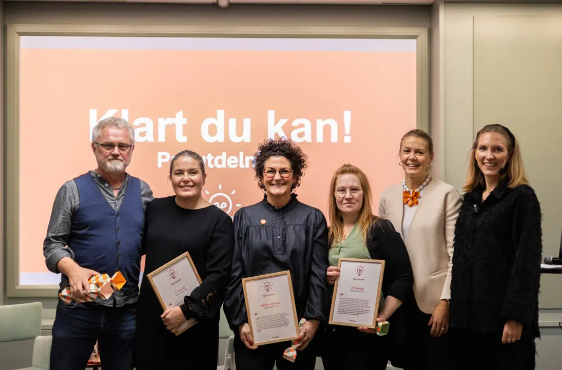 Grupp människor vid en prisutdelning, håller certifikat framför en skylt med texten "Klart du kan!" i en konferensrumsmiljö
