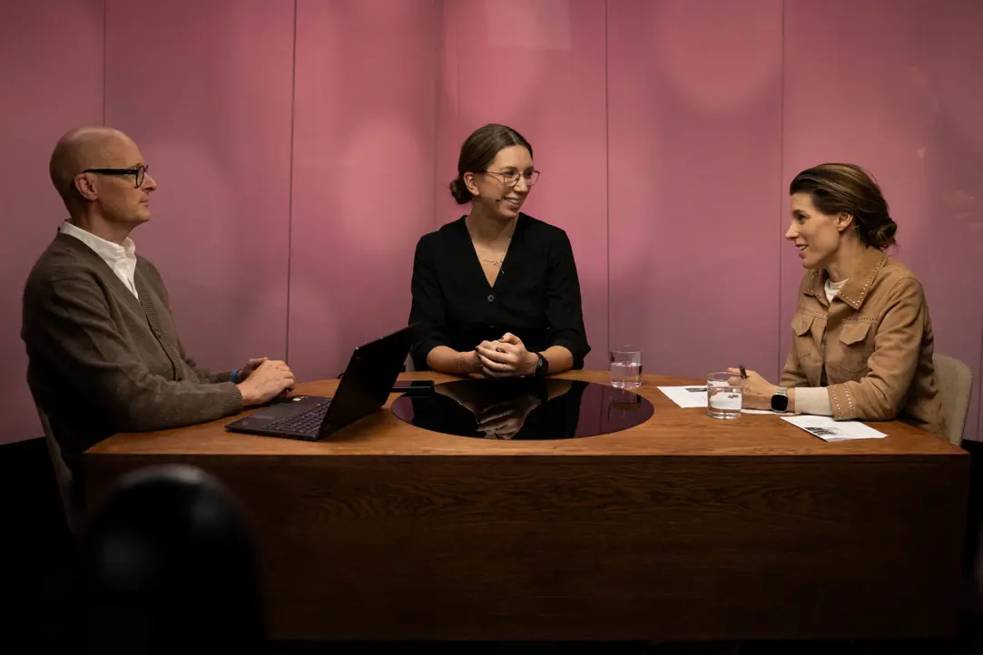 Tre personer deltar i en diskussion vid ett bord med en bärbar dator och glas vatten, med en rosa bakgrund