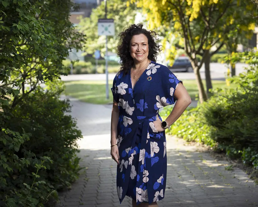 Kvinna i blå klänning med blommönster står på en solig parkväg omgiven av grönska