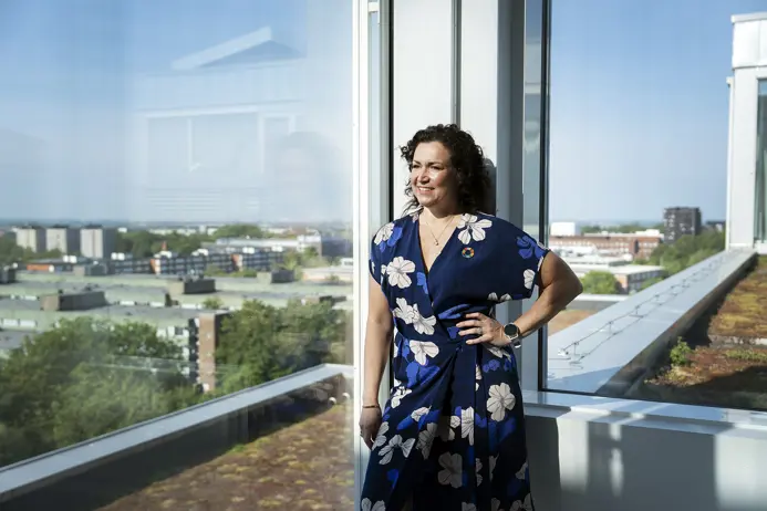 Kvinna i blå klänning med blommönster står vid fönster med utsikt över stad och grönska