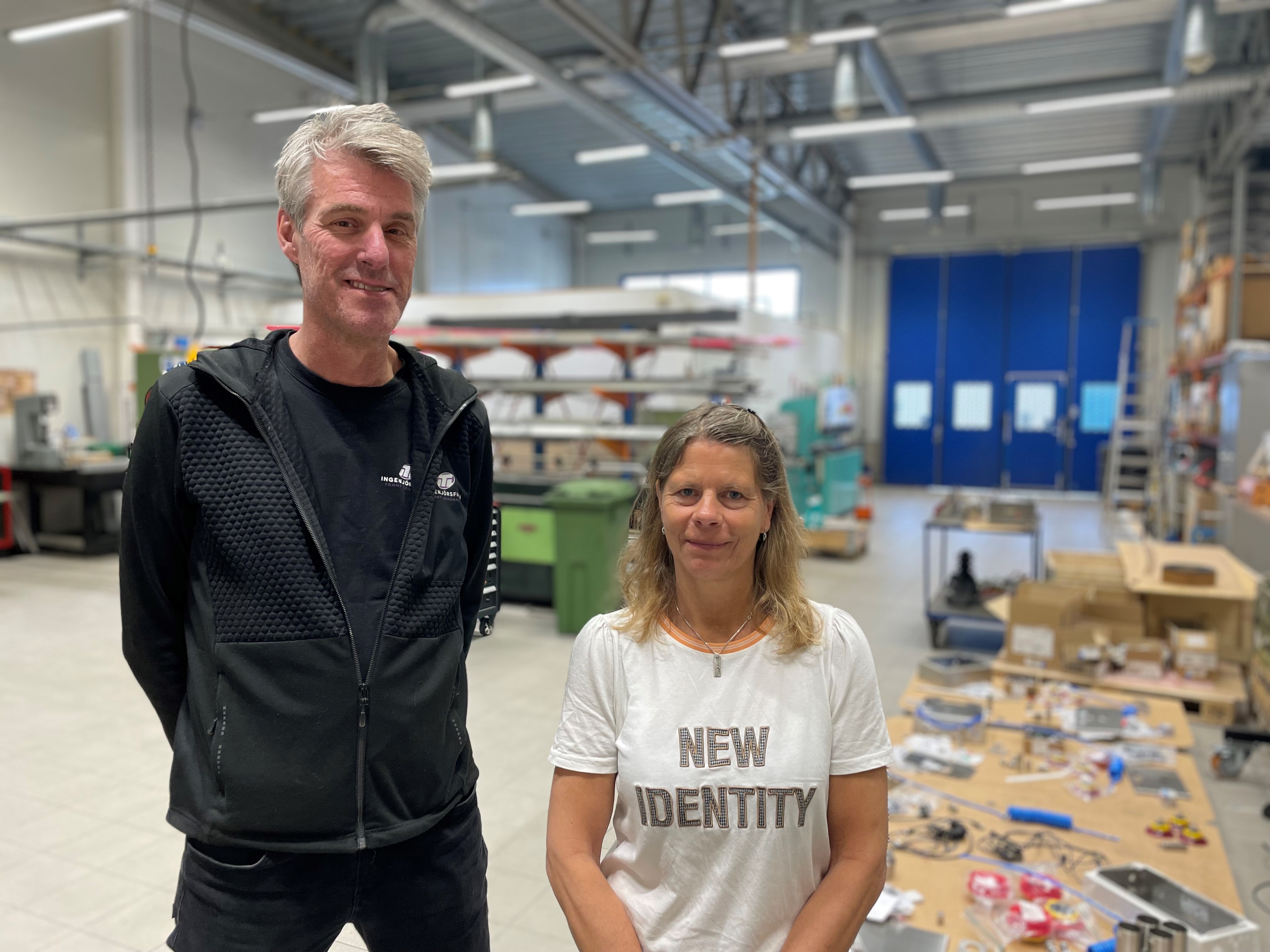 Två personer i en verkstadsmiljö med maskiner och verktyg i bakgrunden. Personen till vänster bär en svart jacka och personen till höger har en vit t-shirt med texten "New Identity"