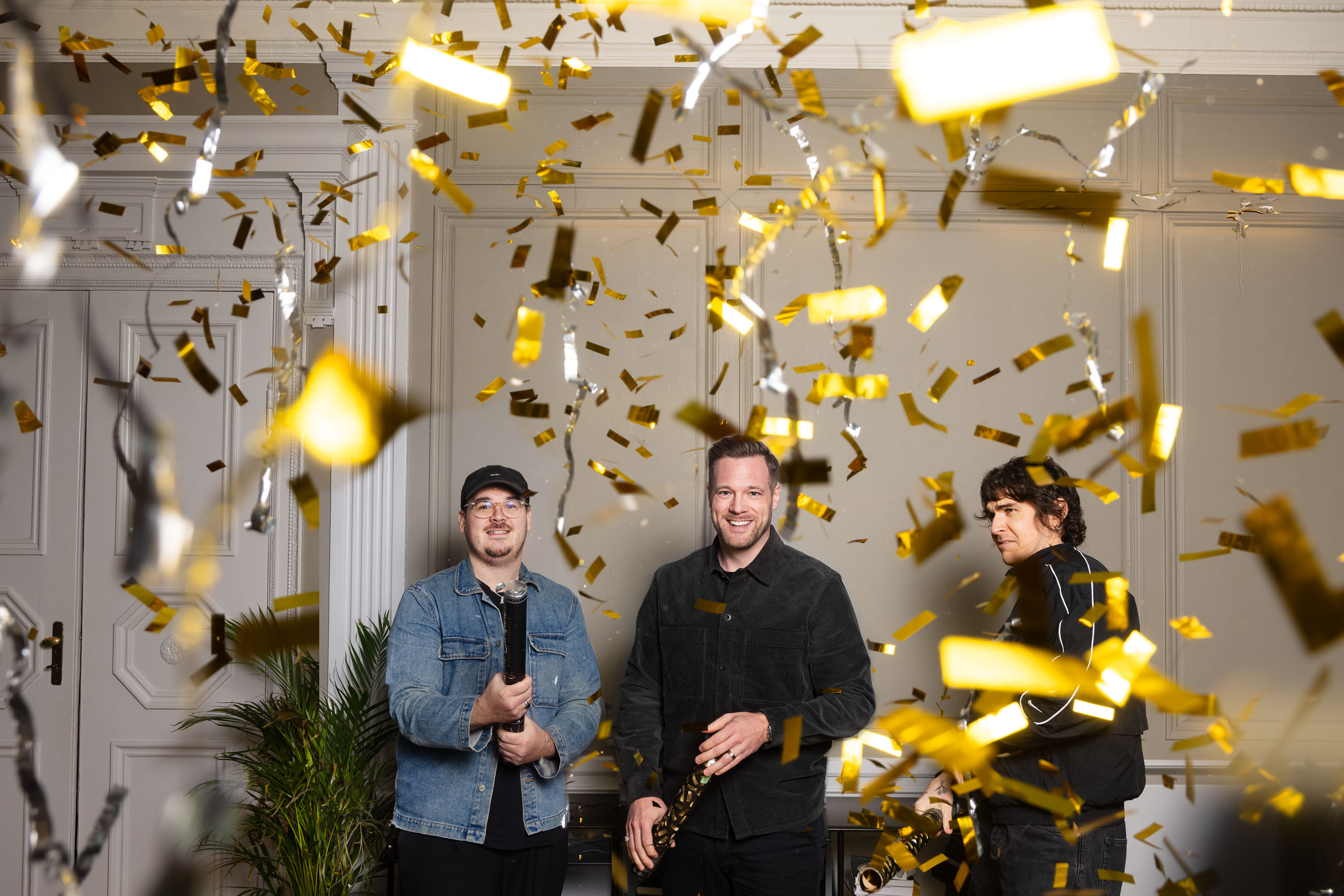 Tre personer firar med konfetti i ett vitt rum, omgivna av växter. Två av dem håller konfettirör medan guldremsor faller från taket. Stämningen är festlig och glad