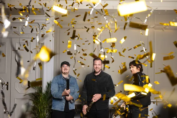 Tre personer firar med konfetti i ett vitt rum, omgivna av växter. Två av dem håller konfettirör medan guldremsor faller från taket. Stämningen är festlig och glad