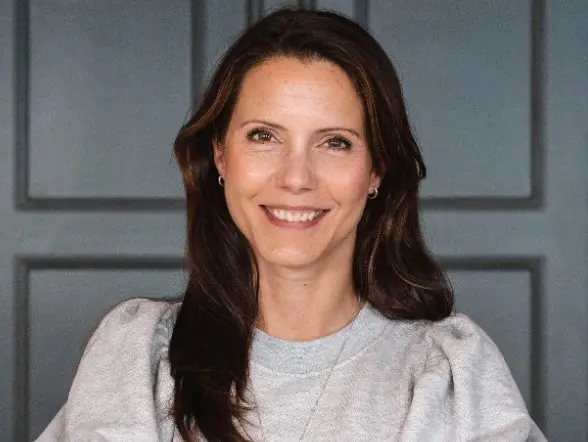 Kvinna med långt mörkt hår ler mot kameran, iklädd en grå tröja framför en mörk bakgrund