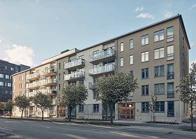 Flervåningshus med modern design och balkonger, omgiven av träd längs en stadsgata i Sverige