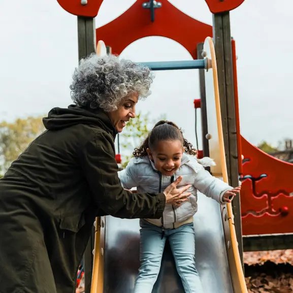 Äldre kvinna hjälper barn att åka rutschkana på lekplats