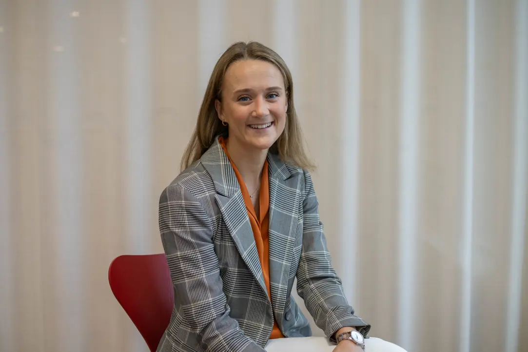 Person i grå-rutig kavaj och orange topp sitter på en röd stol, med en ljus gardinbakgrund och ler mot kameran