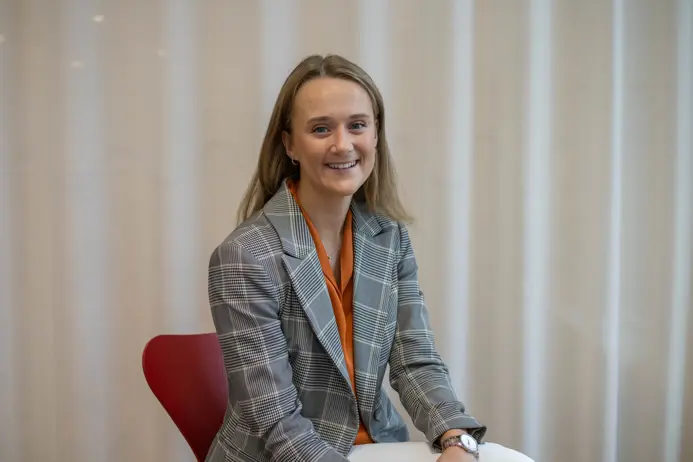 Person i grå-rutig kavaj och orange topp sitter på en röd stol, med en ljus gardinbakgrund och ler mot kameran