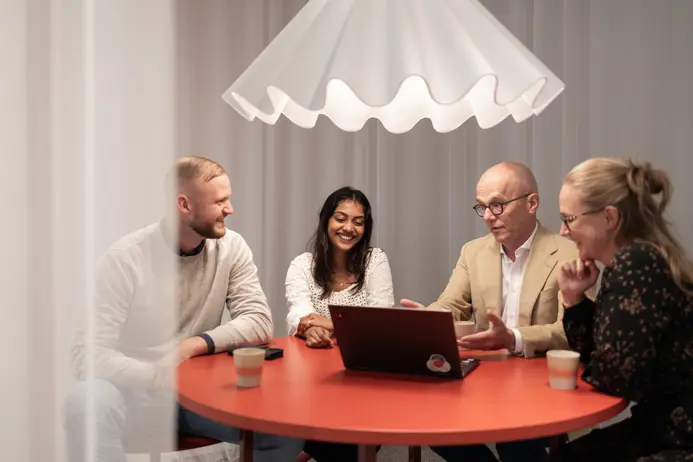 Fyra personer har ett affärsmöte runt ett runt, rött bord under en vit lampa. De diskuterar något med hjälp av en bärbar dator