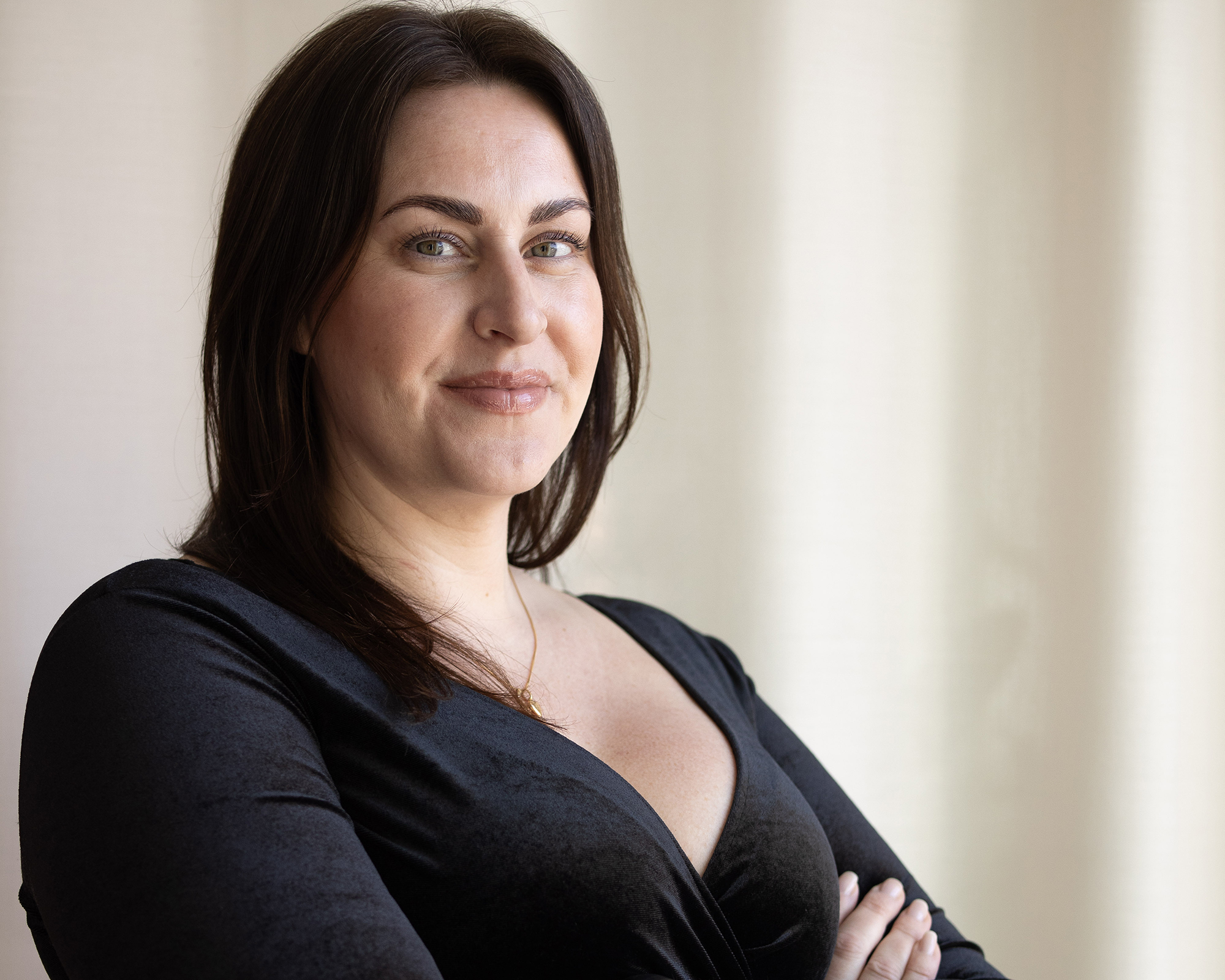 Kvinna med mörkt hår och svart klänning står med armarna i kors och ler mot kameran, mot en ljus bakgrund