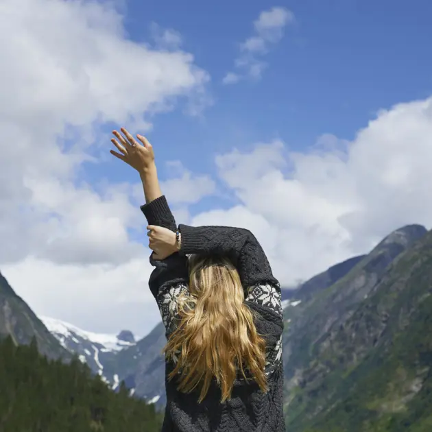 Kvinna i stickad tröja sträcker sig mot himlen i en naturskön bergsmiljö med snöklädda toppar och klarblå himmel