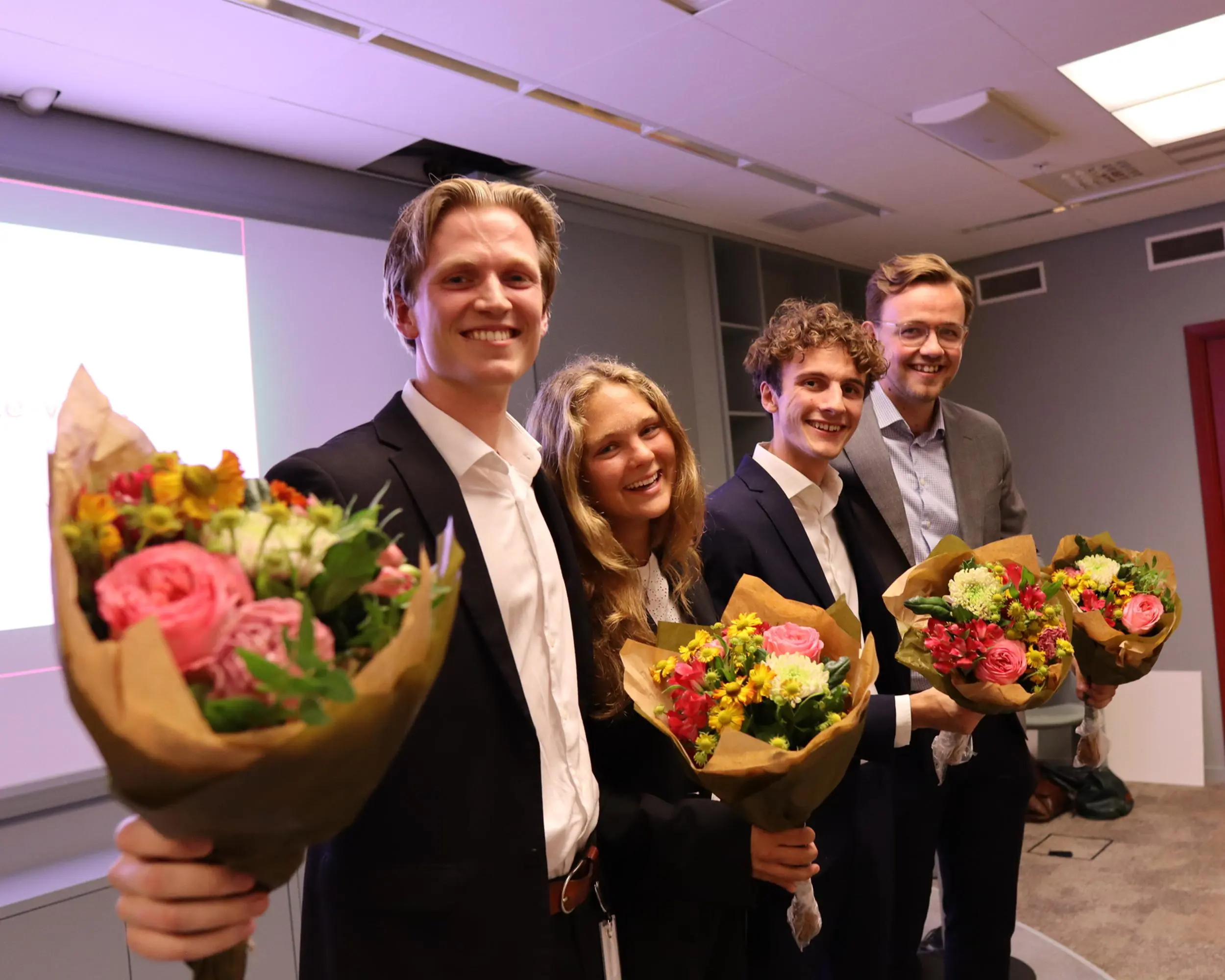 Fyra personer i kostym står tillsammans och håller färgglada blombuketter i ett konferensrum. De verkar glada och nyss ha deltagit i en presentation eller ett evenemang. Bakgrunden visar en presentation och en modern inredning