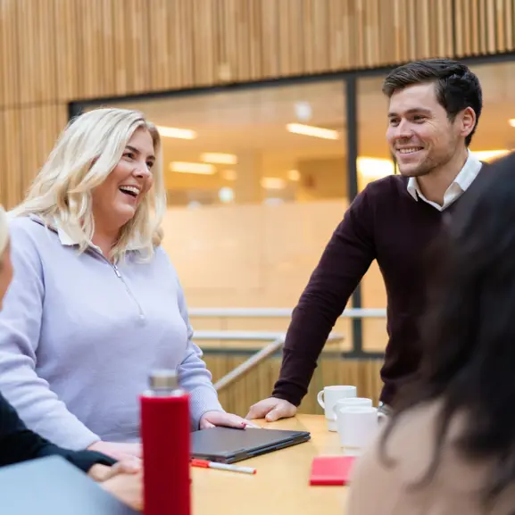 Två leende människor i ett modernt kontorsmiljö, som diskuterar och samarbetar vid ett bord med bärbar dator och kaffekoppar