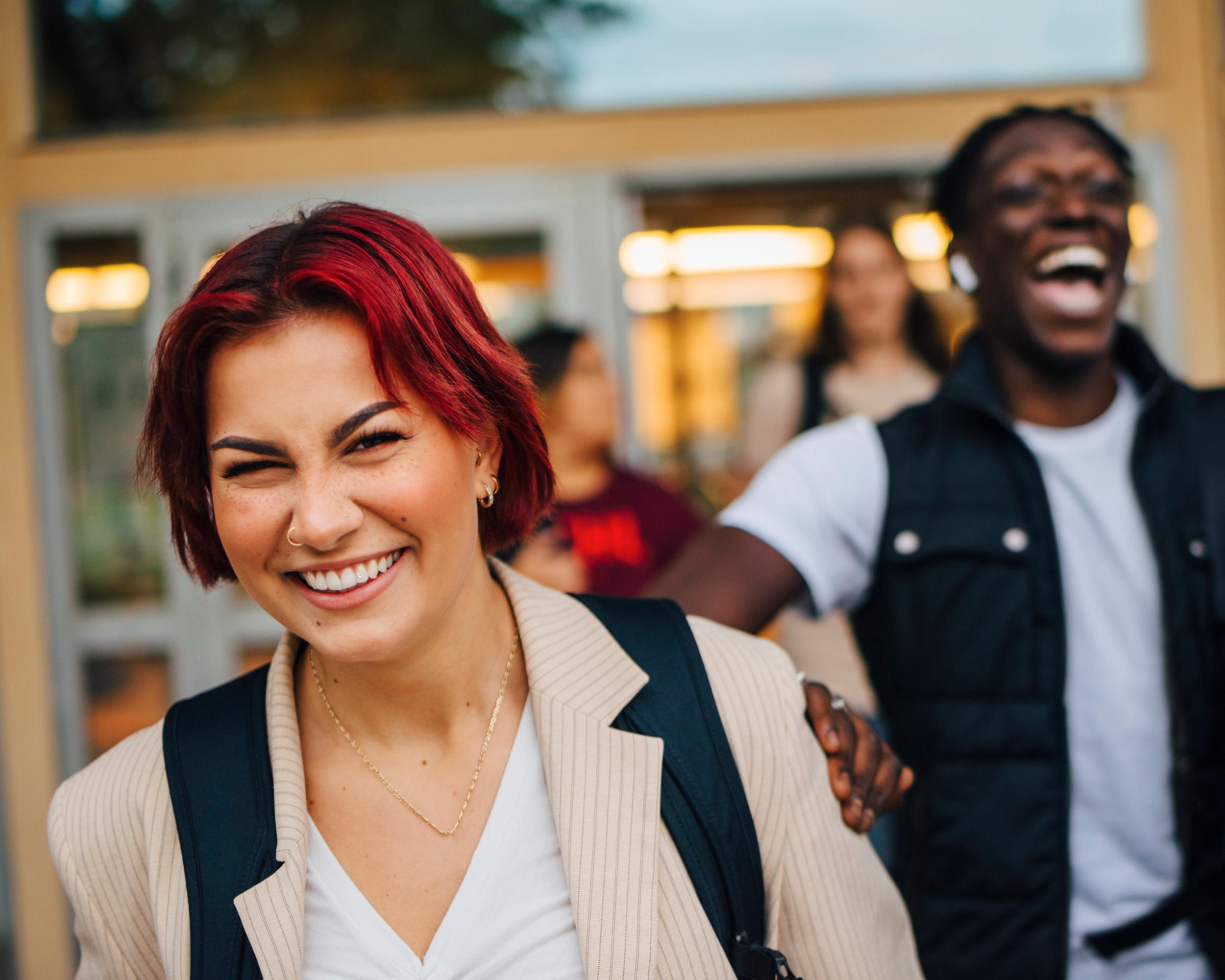 Glada studenter utanför skolbyggnad, en med rött hår och en med svart väst, skrattar tillsammans