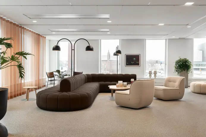 Modern kontorsmiljö med stilrena, jordnära färgtoner. En brun, modulär soffa och två beige fåtöljer omger ett litet runt bord i centrum. Stora fönster erbjuder rikligt med dagsljus och en utsikt över staden. Gröna växter och dekorativa detaljer ger rummet en avslappnad atmosfär