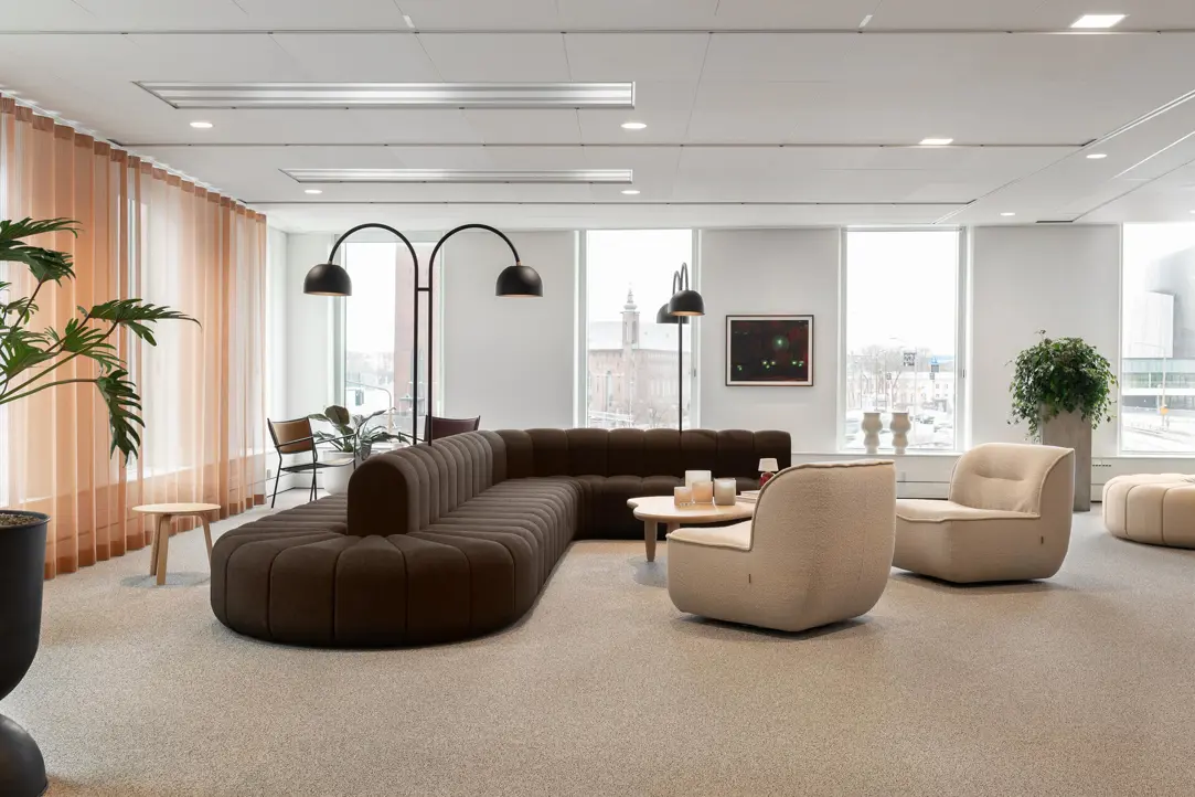 Modern kontorsmiljö med stilrena, jordnära färgtoner. En brun, modulär soffa och två beige fåtöljer omger ett litet runt bord i centrum. Stora fönster erbjuder rikligt med dagsljus och en utsikt över staden. Gröna växter och dekorativa detaljer ger rummet en avslappnad atmosfär