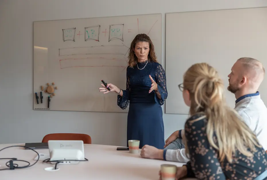 Kvinna ger presentation för två personer vid bord, med whiteboard i bakgrunden