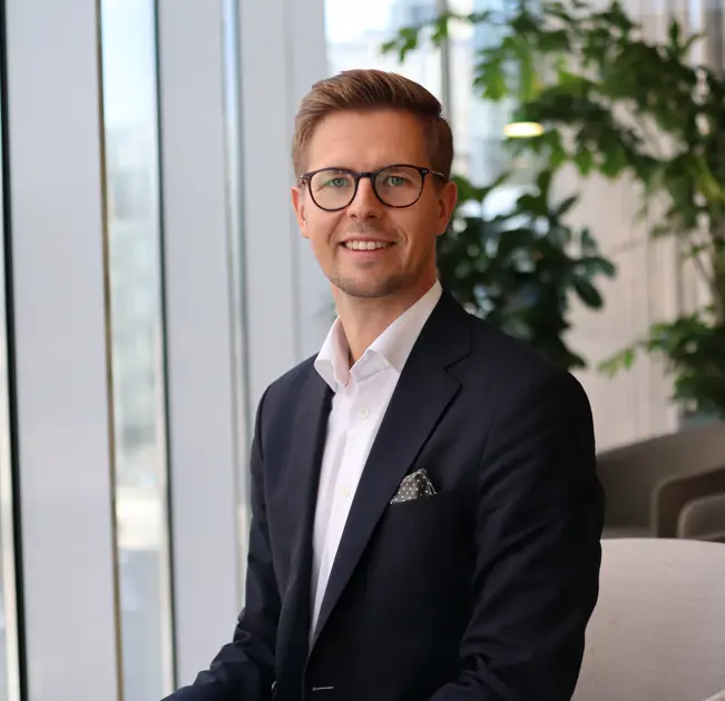 Man i kostym och glasögon sitter i en modern kontorsmiljö med stora fönster och gröna växter i bakgrunden