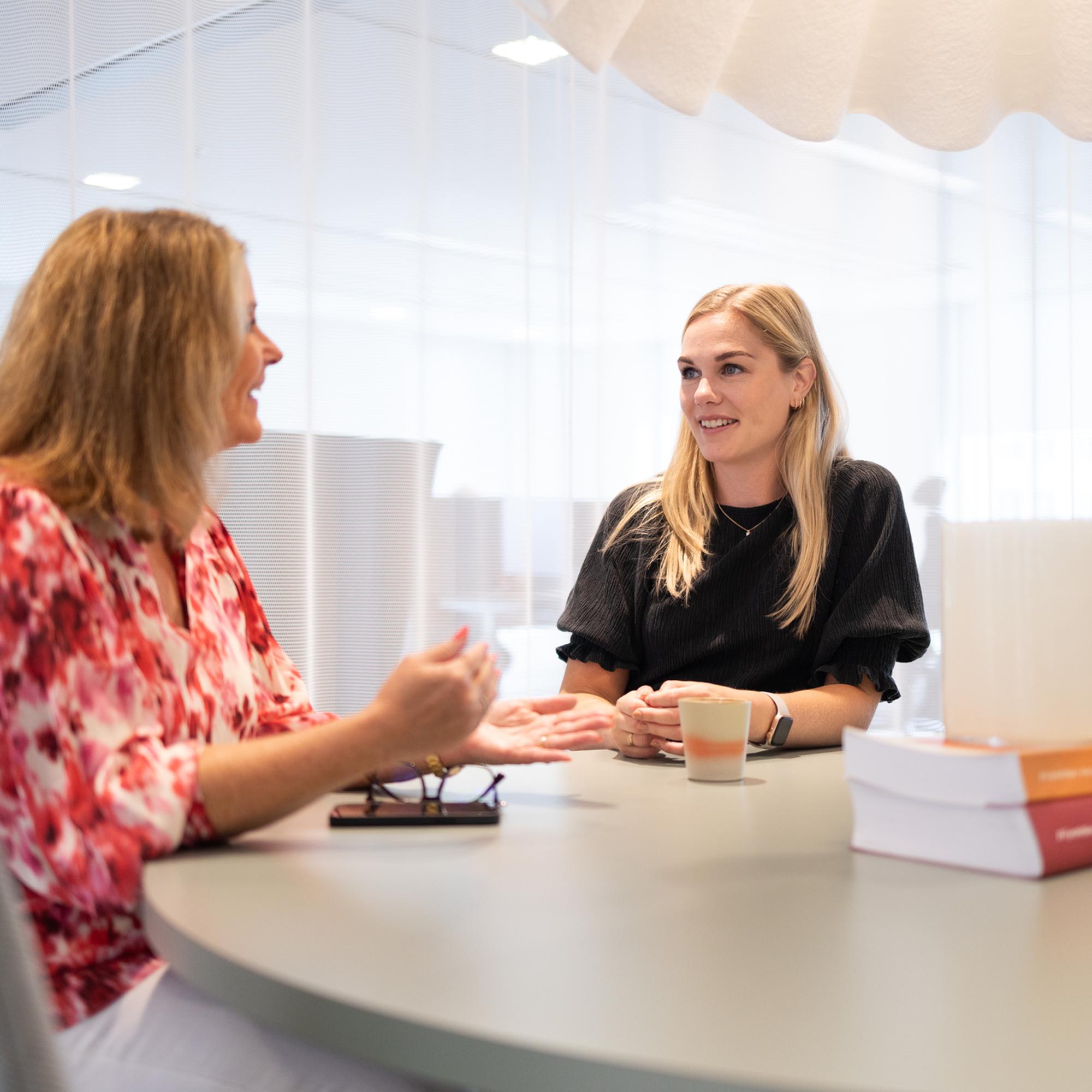 Två kvinnor diskuterar vid ett bord på kontoret, en bär en röd blus och den andra en svart topp. De verkar vara engagerade i en avslappnad konversation under ett möte, med kaffekopp och böcker närvarande på bordet