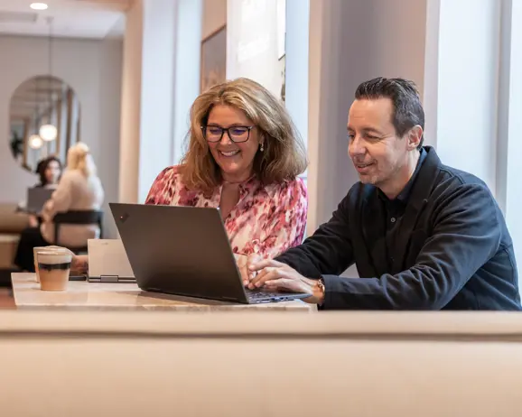 Två glada personer arbetar tillsammans vid en laptop på ett café, med kaffe på bordet och andra människor i bakgrunden
