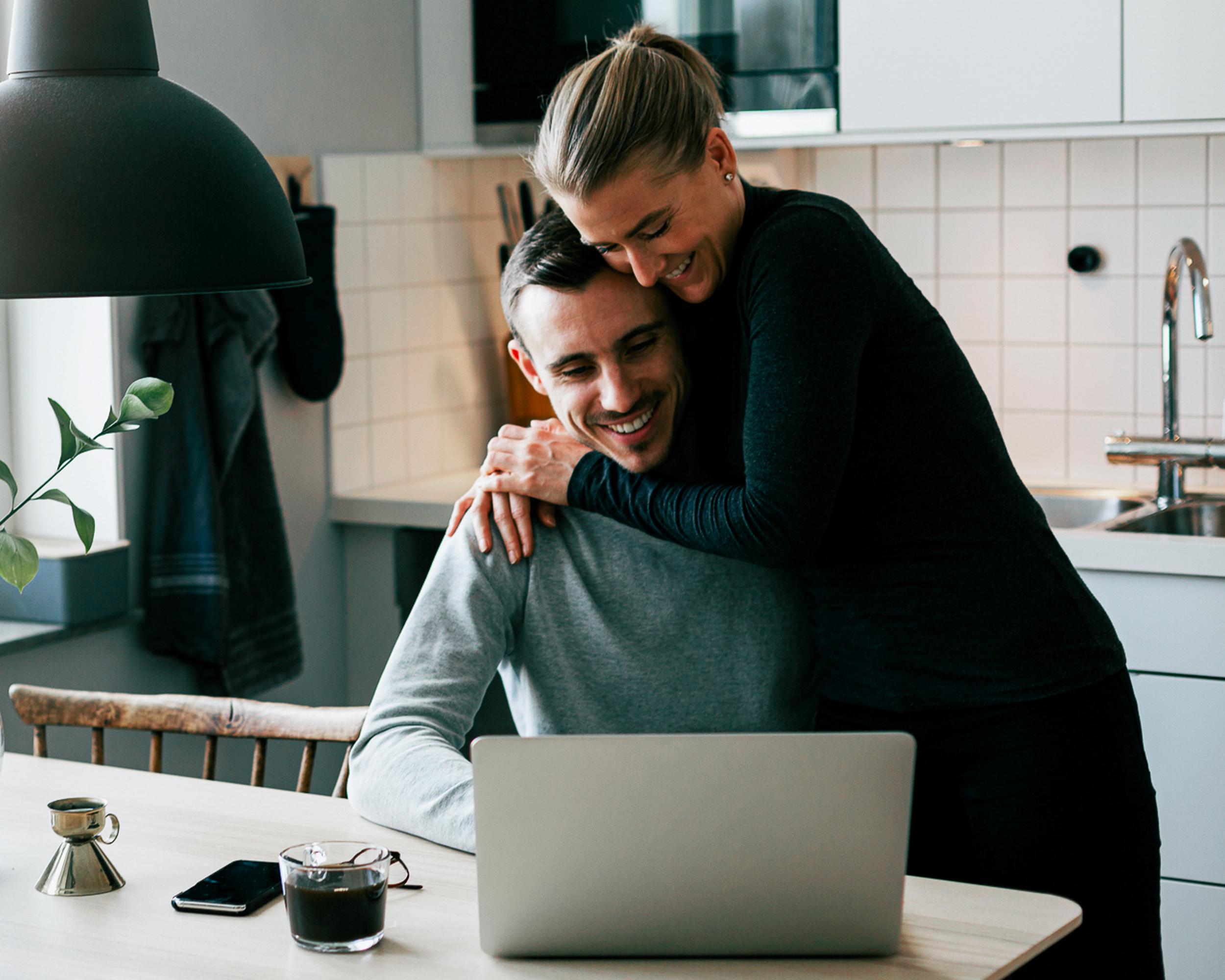 Par i köket som ler och använder en bärbar dator, med en kopp kaffe på bordet