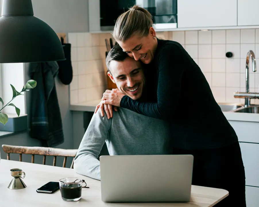 Par i köket som ler och använder en bärbar dator, med en kopp kaffe på bordet