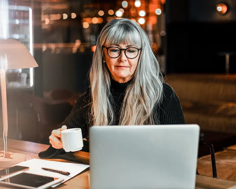 Äldre kvinna med långt grått hår och glasögon arbetar vid en bärbar dator medan hon håller i en kaffekopp