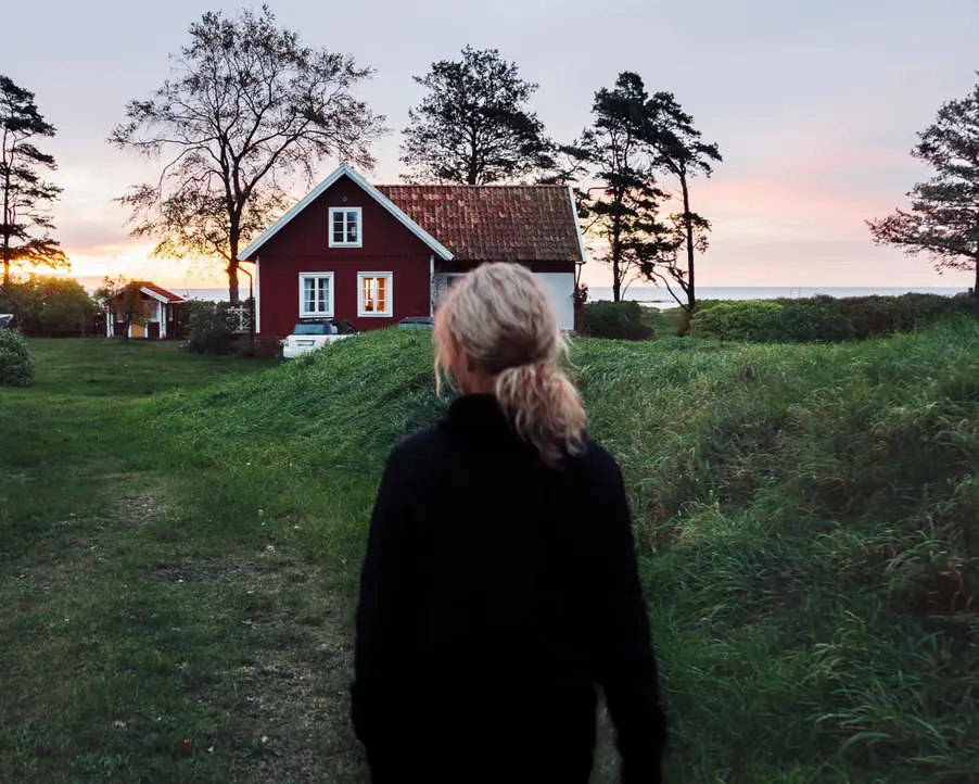 Kvinna går mot en röd stuga på landsbygden vid solnedgång, omgiven av grönska och träd nära havet