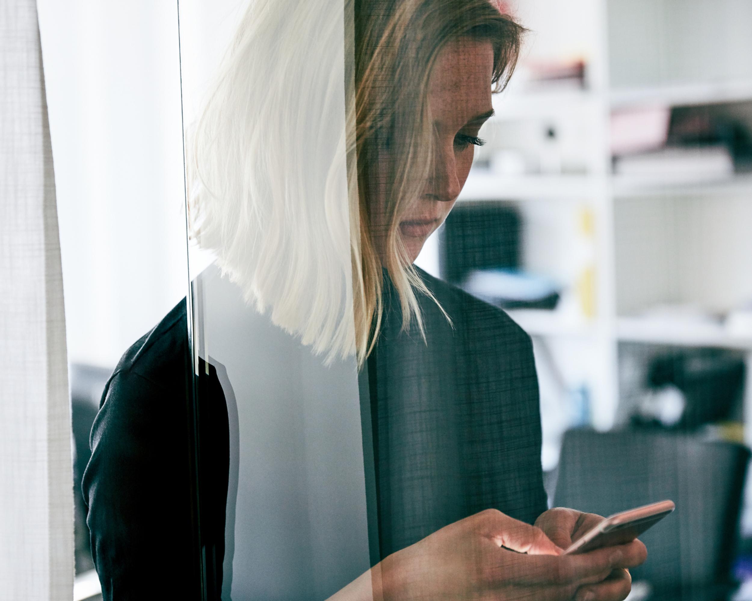 Kvinna med blont hår som använder smartphone bakom glasvägg i kontorsmiljö