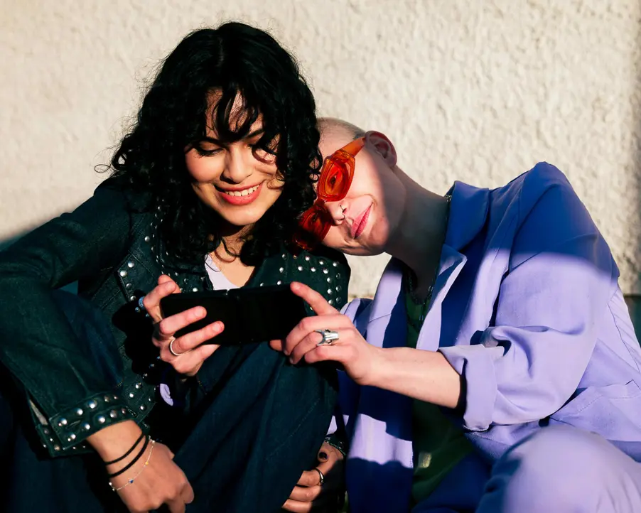 Två leende personer sitter nära varandra och tittar på en mobiltelefon utomhus i soligt väder. En person bär en lila jacka och glasögon, medan den andra har lockigt hår och en denimjacka