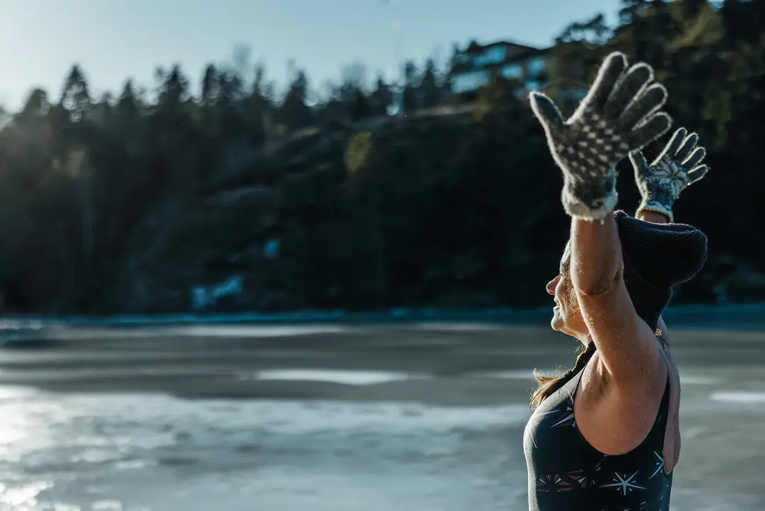 Kvinna som njuter utomhus, klädd i baddräkt och vinterhandskar, med händerna upp i luften vid en isig sjö