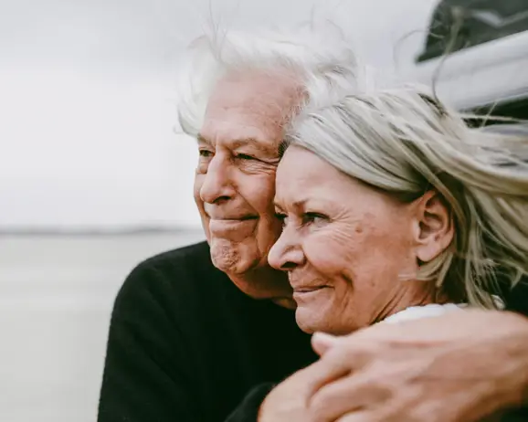 Äldre par omfamnar varandra vid stranden, ser glada och harmoniska ut