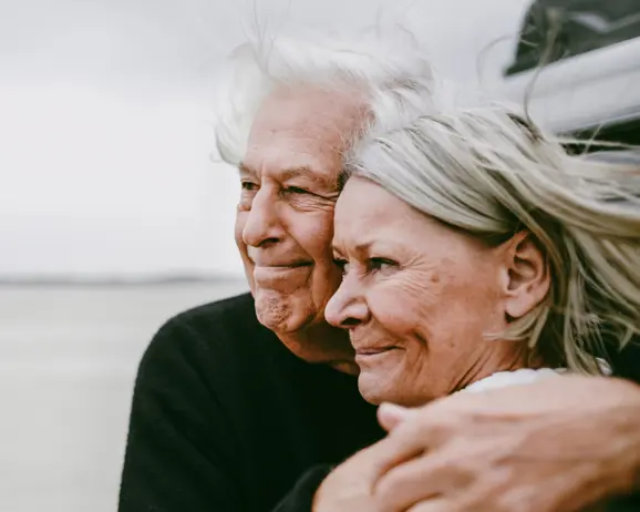 Äldre par omfamnar varandra vid stranden, ser glada och harmoniska ut