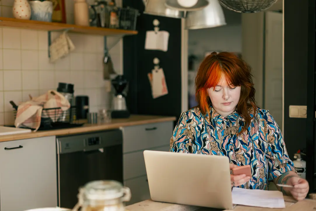 Kvinna med rött hår arbetar vid en laptop i ett modernt kök med rostfri diskbänk och trähyllor
