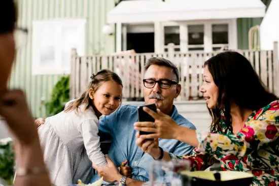 Familj som sitter utomhus och använder en smartphone tillsammans, med ett barn i fokus och ett grönt hus i bakgrunden