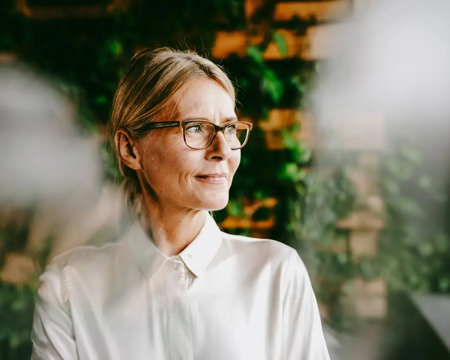 Kvinna med glasögon i vit skjorta tittar bortåt framför en grön växtbakgrund
