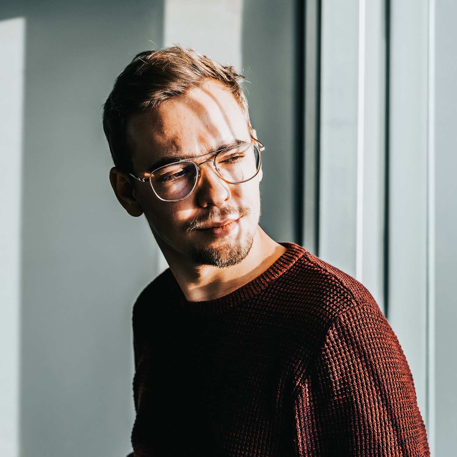 Man med glasögon i dagsljus, bär en stickad tröja, står vid fönstret