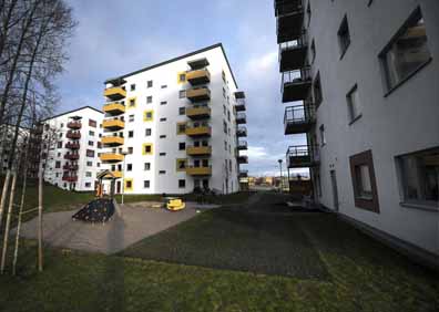 Flerfamiljshus med vita fasader och gula balkonger i en stadsdel, omgiven av gröna gräsmattor och lekplats