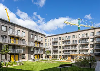 Modern bostadsområde med lägenhetsbyggnader och gröna innergårdar under blå himmel, byggkranar i bakgrunden