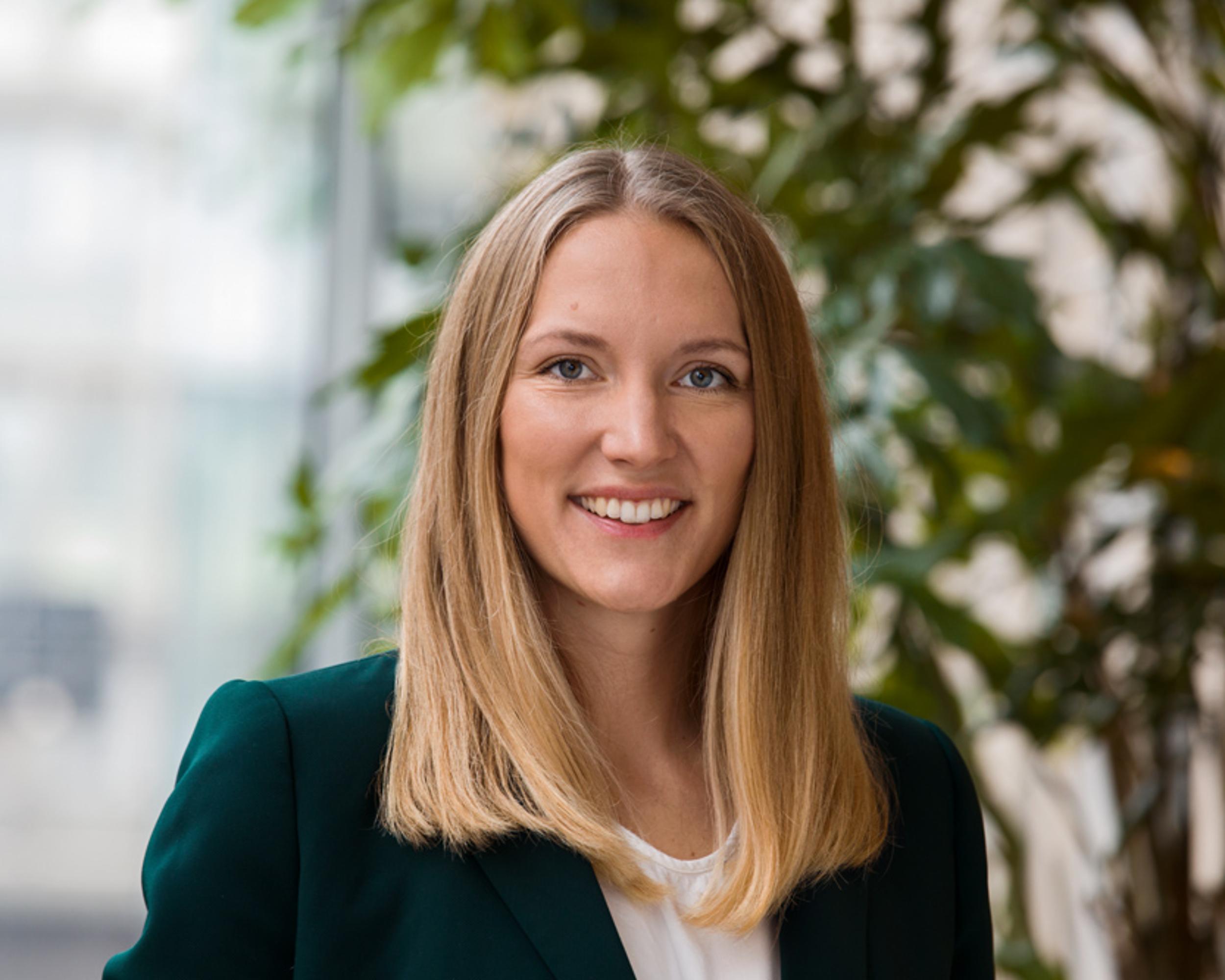 Kvinna med blont hår i grön kavaj, leende mot kameran, med grönskande växt i bakgrunden