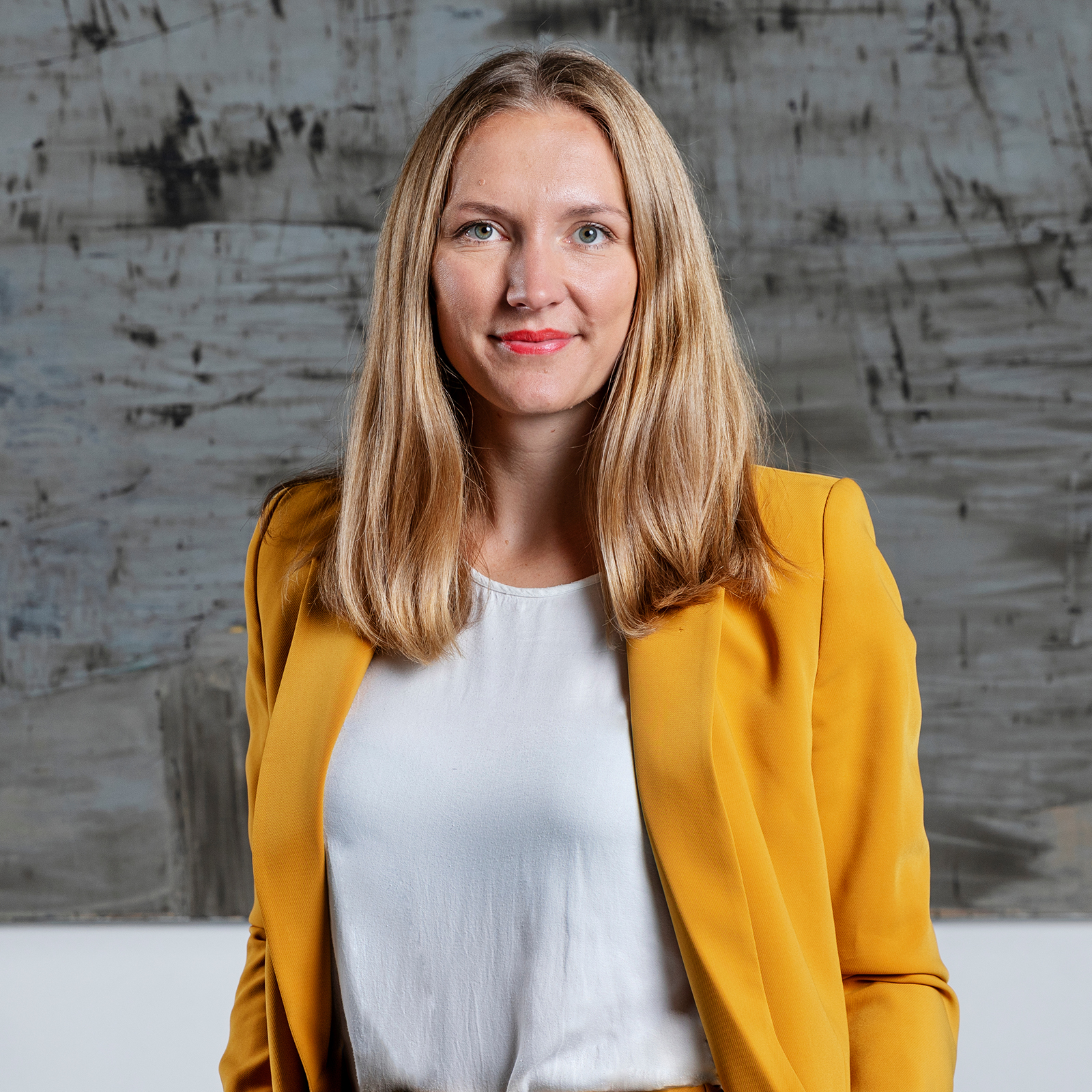 Kvinna med långt blont hår i gul kavaj och vit blus står framför en abstrakt grå bakgrund