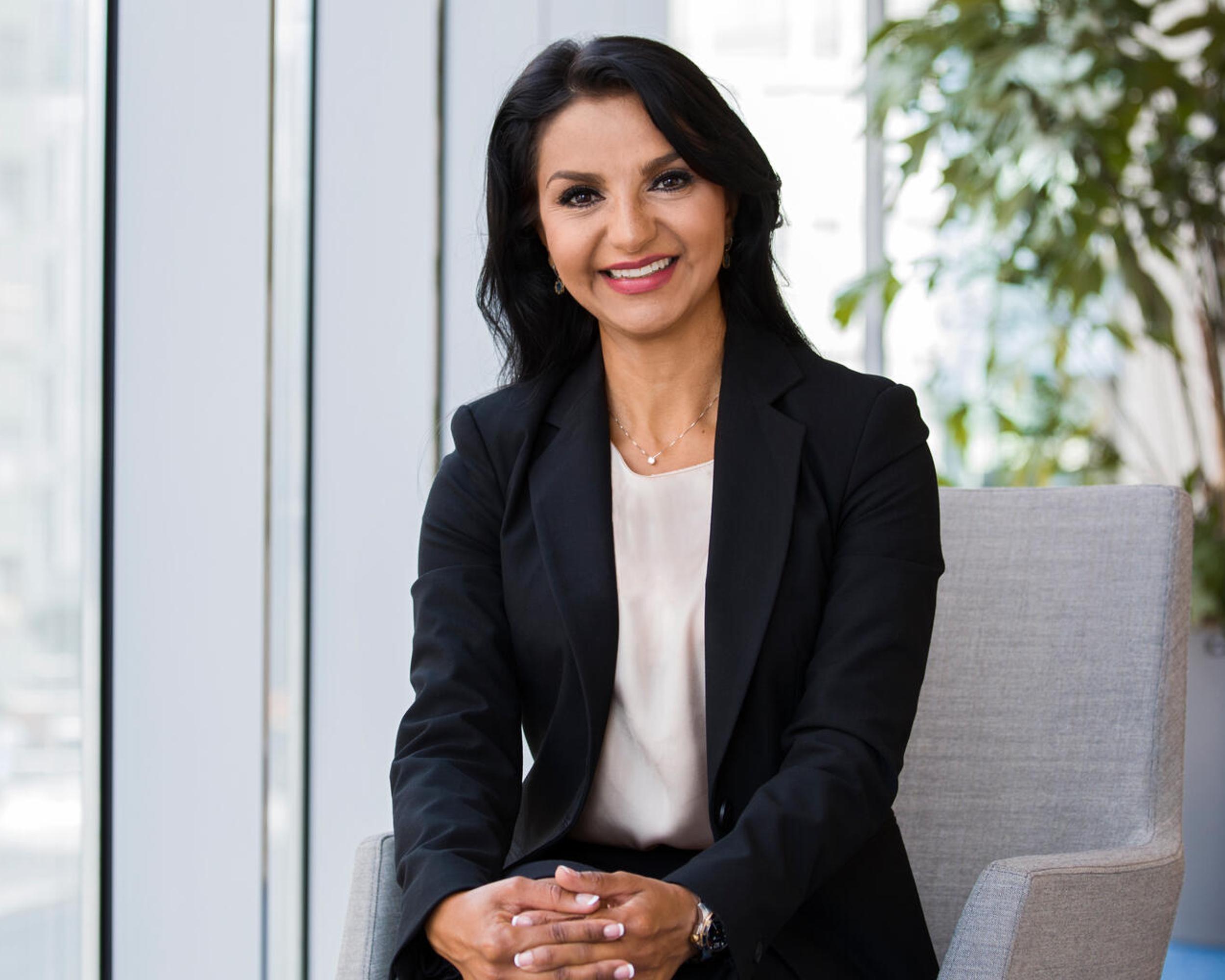 Kvinna i affärskläder sitter leende på en stol i ett kontor med stora fönster och gröna växter i bakgrunden